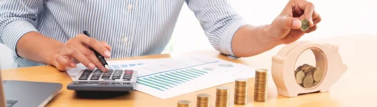 Person calculating finances with coins and charts to compare savings account vs checking account options at First National Bank of Brookfield in Brookfield, Illinois.